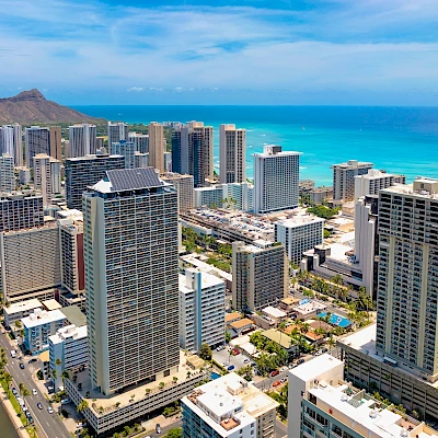 An aerial view of a coastal city with tall buildings, a blue ocean, and a mountain in the background under clear skies.