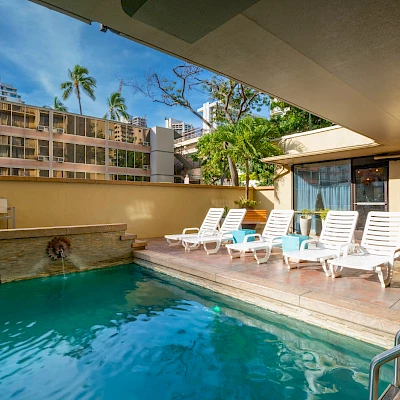 The image shows an outdoor swimming pool with sun loungers, surrounded by buildings and trees under a blue sky.