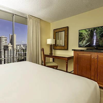 A cozy hotel room with a large bed, city view, TV on a wooden cabinet, desk, lamp, mirror, and sliding door leading to a balcony ending the sentence.