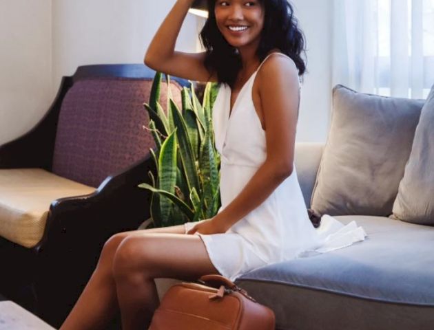 A woman in a white dress sits on a sofa, smiling, holding sunglasses, with a brown backpack by her side.
