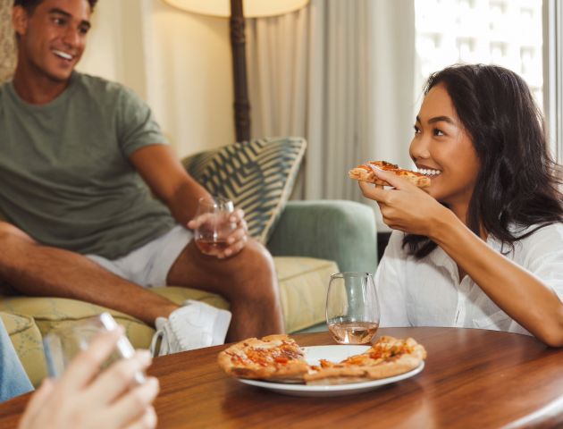 A group of people enjoying pizza and drinks in a cozy living room setting, smiling and engaging in conversation.
