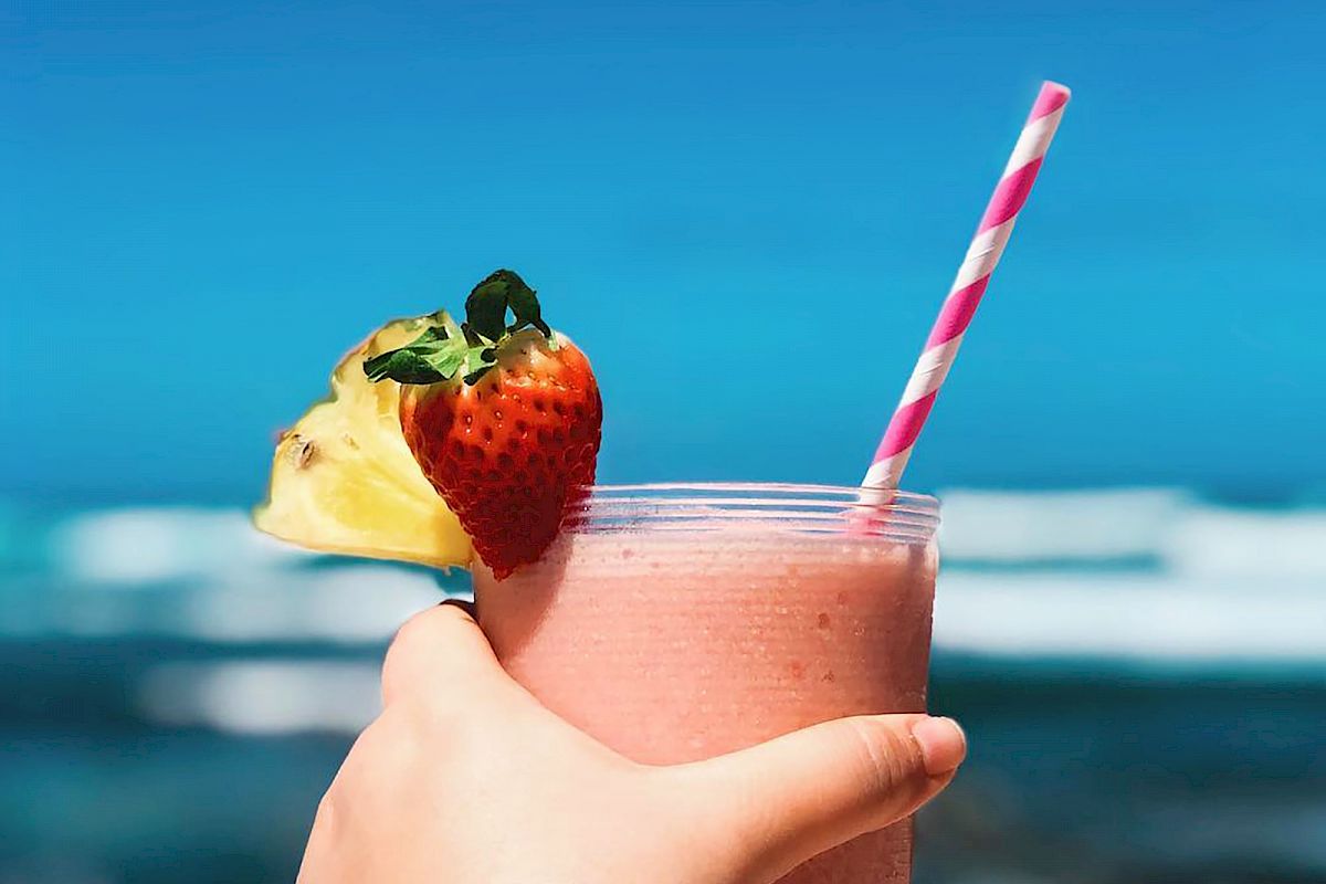 A hand holding a pink smoothie with a striped straw, topped with a strawberry and a pineapple slice, against a backdrop of a beach and ocean.