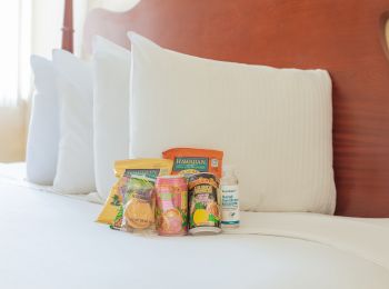 The image shows snack items and a bottle of water arranged on a white bed with pillows and a wooden headboard in the background.