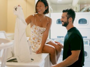 A man and woman are in a laundry room, with the woman sitting on a washer and holding up a piece of clothing while the man stands nearby, smiling.
