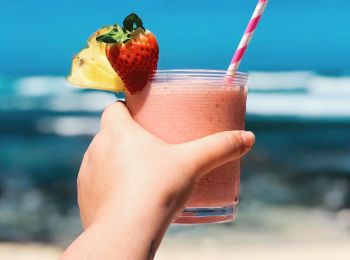 A hand holds a pink smoothie with a striped straw, garnished with a strawberry and pineapple slice, against a beach backdrop.