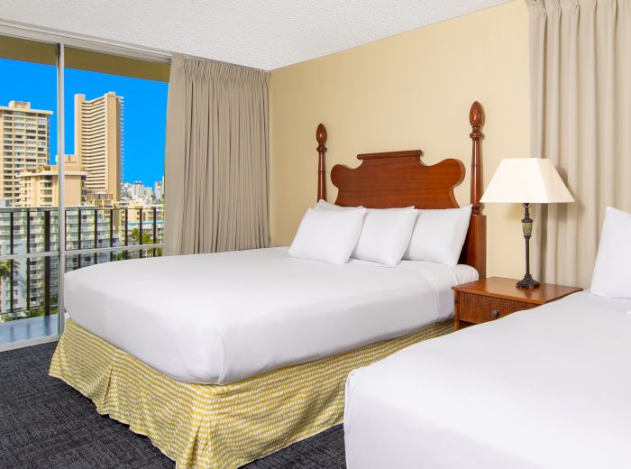 A hotel room with two beds, side table with lamp, beige curtains, and a cityscape view from a large window.
