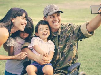 A happy family, including a person in military uniform and a wheelchair, is taking a selfie together in an outdoor setting.