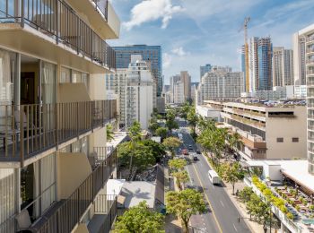 Urban street view with tall buildings, balconies, parked cars, and trees under a partly cloudy sky.