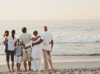 A group of six people, including children and adults, stand on a beach with their backs to the camera, facing the ocean together.