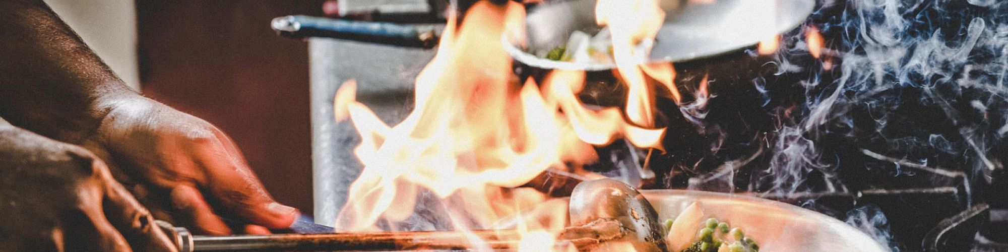 A chef is cooking with intense flames and smoke in a busy kitchen, using a metal spoon to stir ingredients in a skillet.