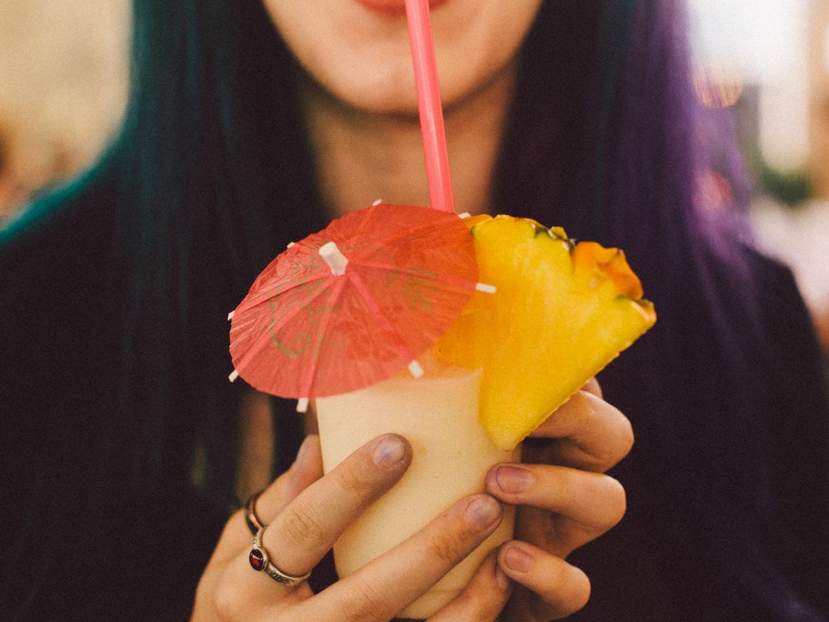 A person holds a tropical drink garnished with pineapple slices and a small umbrella, sipping through a pink straw, with a colorful background.