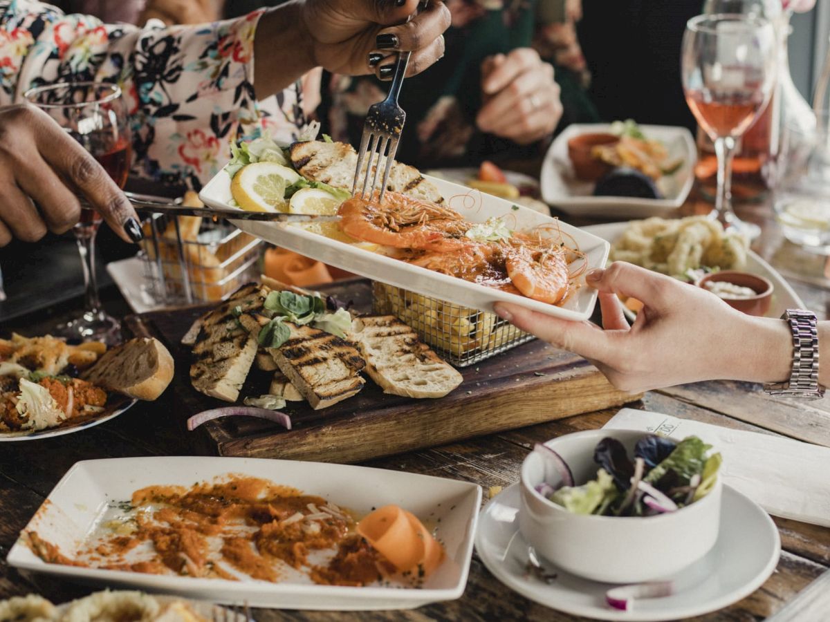 A group of people are sharing a meal, with dishes including grilled shrimp, salad, bread, and wine, all laid out on a wooden table.