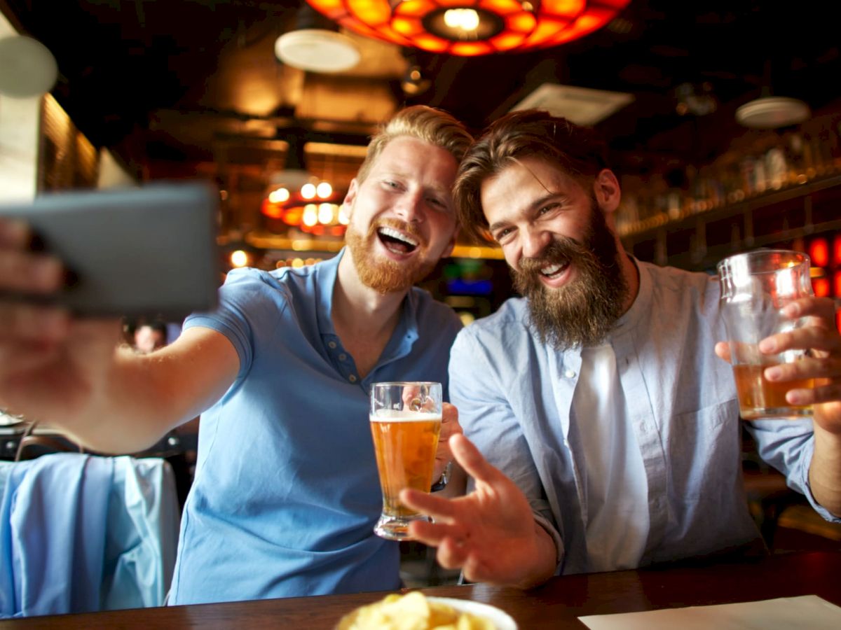 Two men are at a pub, smiling and taking a selfie while holding glasses of beer, with a warm, inviting atmosphere around them.