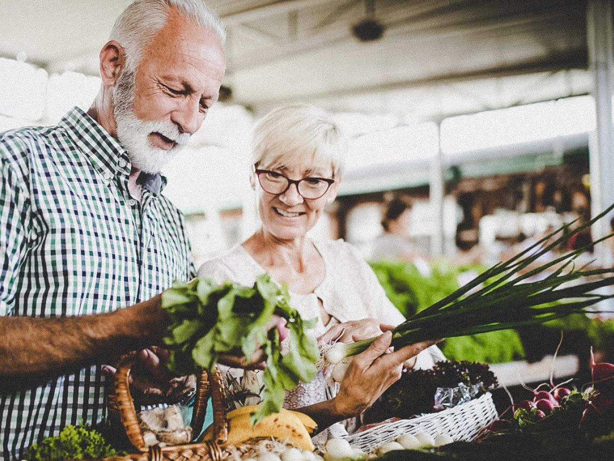 Two elderly individuals are happily selecting fresh vegetables at a market stall, surrounded by various greens and other produce.