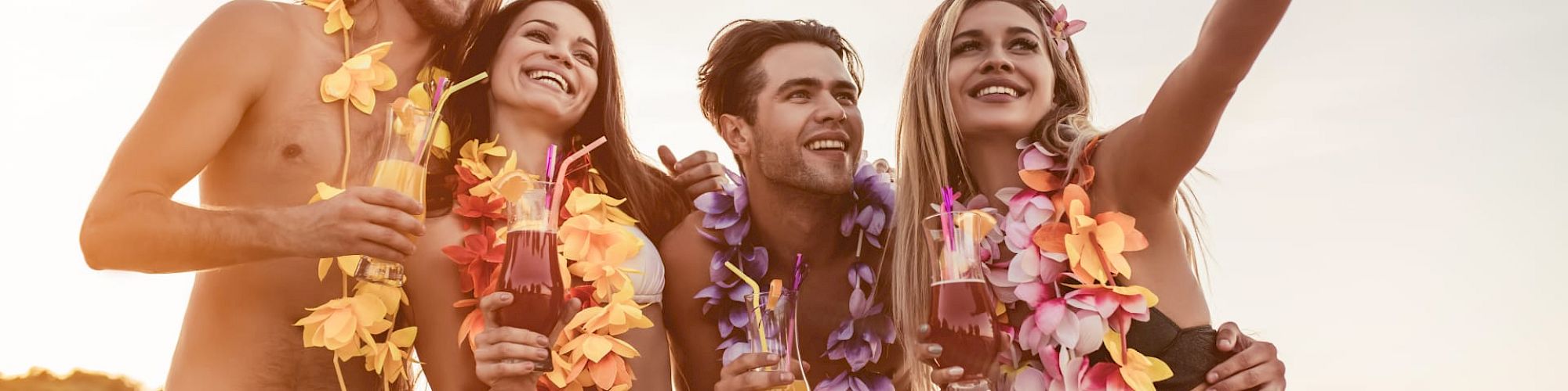 A group of four people, dressed in tropical attire with leis, are taking a selfie on a beach at sunset, holding drinks and smiling.