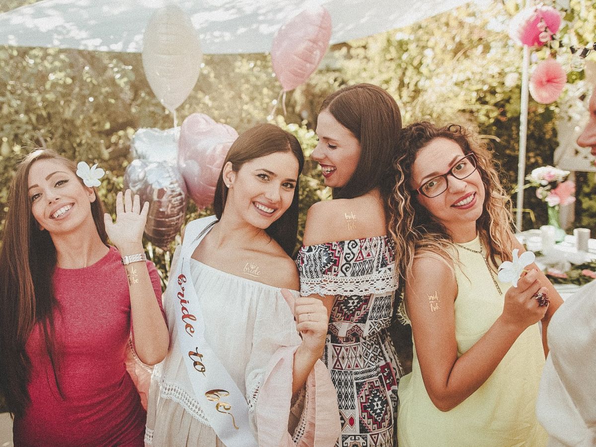 Four women celebrating outdoors with balloons and flowers, smiling and cheerful, one wearing a "bride to be" sash, enjoying a festive event.