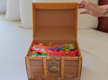 A hand reaches towards an open faux treasure chest filled with colorful children's toys, placed on a white surface.