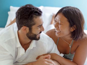 A couple is lying on a bed, facing each other and smiling, creating a warm and intimate moment.