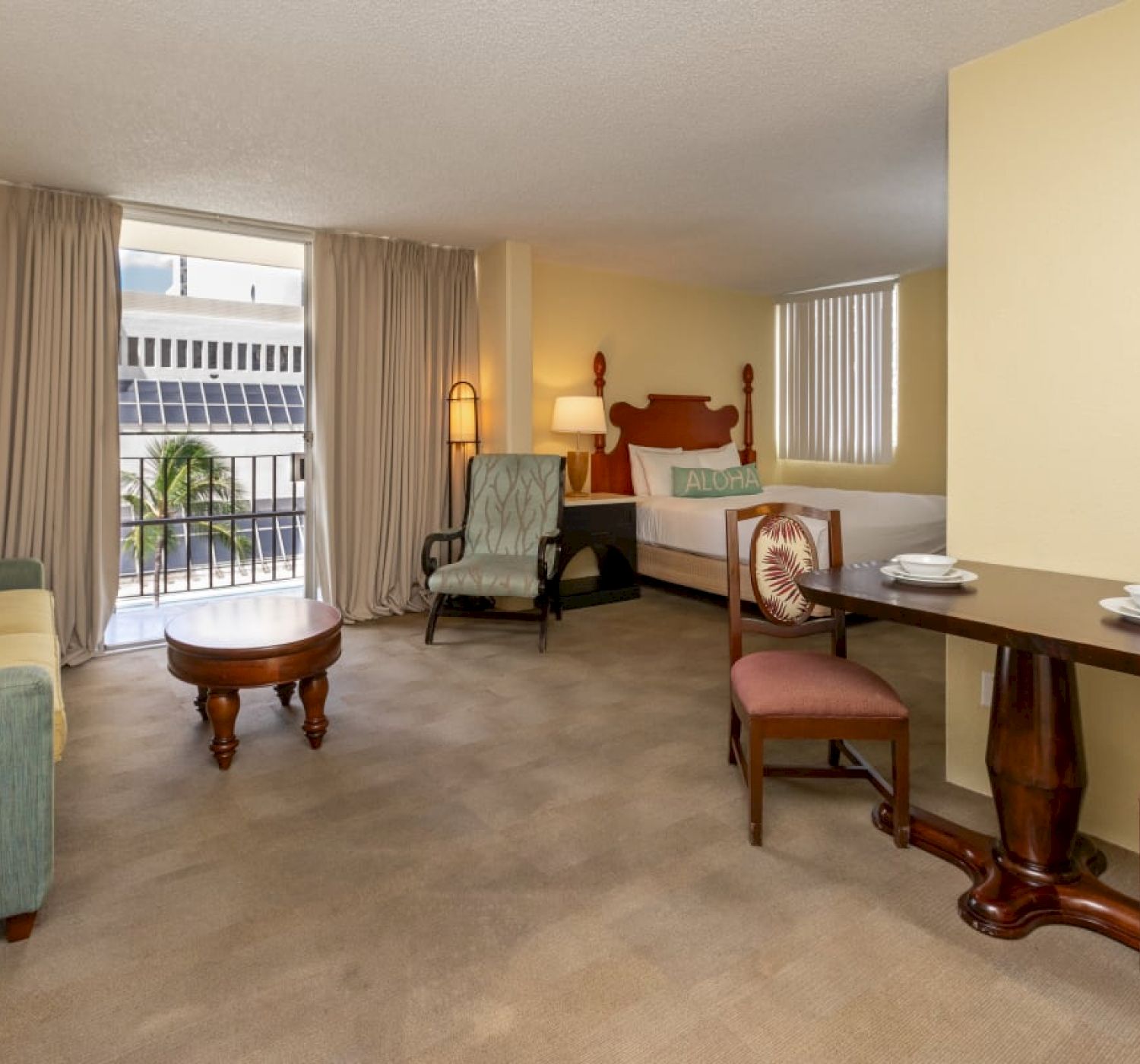 The image shows a hotel room with a seating area, table, chairs, coffee table, bed, and a window with curtains letting in natural light.