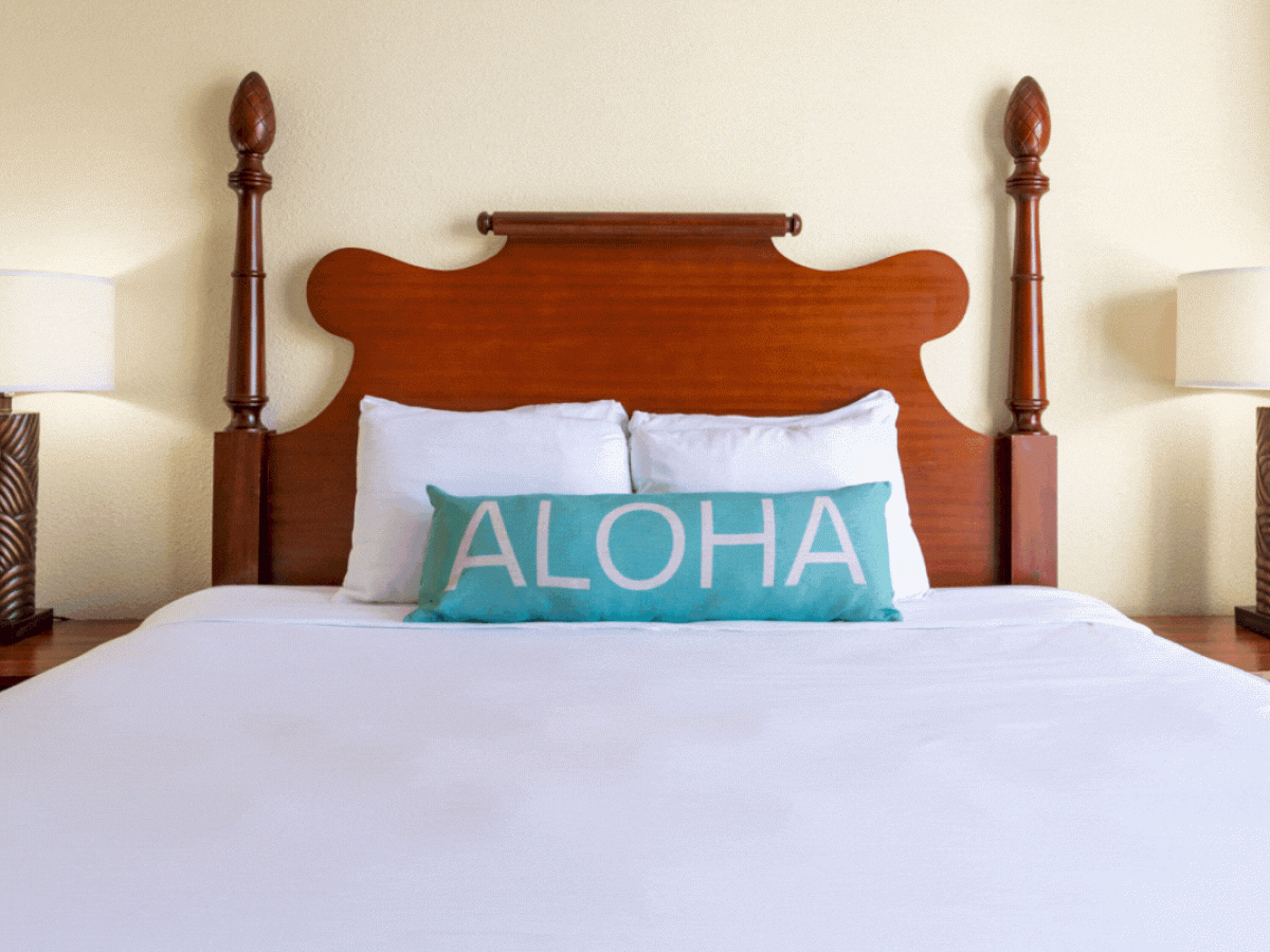 A neatly made bed with a wooden headboard, two pillows, and a decorative pillow with the word "ALOHA." on it, flanked by two bedside lamps.