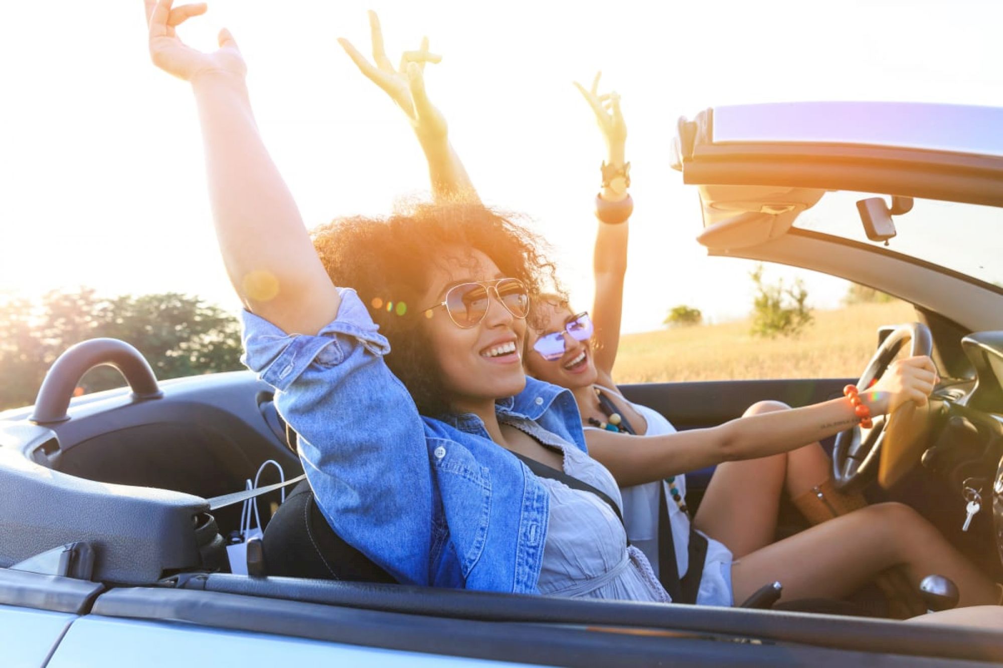 Two people are driving in a convertible with the top down, enjoying the sun with their hands raised and making peace signs.