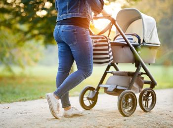 A person is pushing a baby stroller along a path in a park during a sunny day, dressed casually in jeans and a jacket.