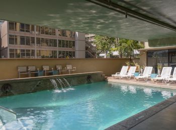The image shows an outdoor pool area with lounge chairs, a small waterfall feature, and buildings in the background under a covered section.